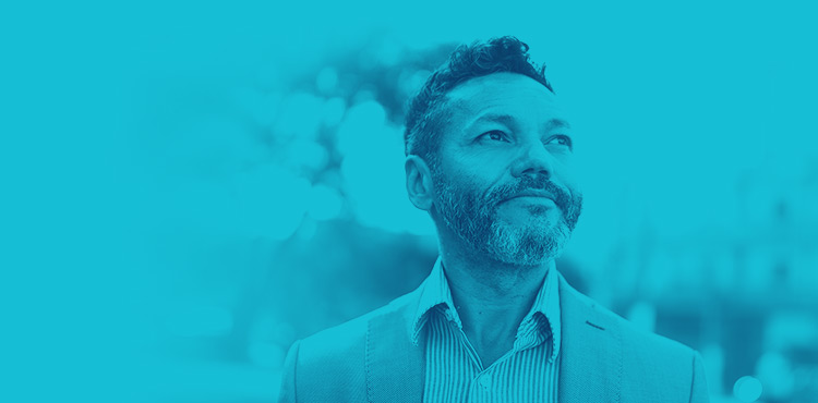A middle-aged man with a scruffy beard and a casual dress coat on looking up and out in the distance.